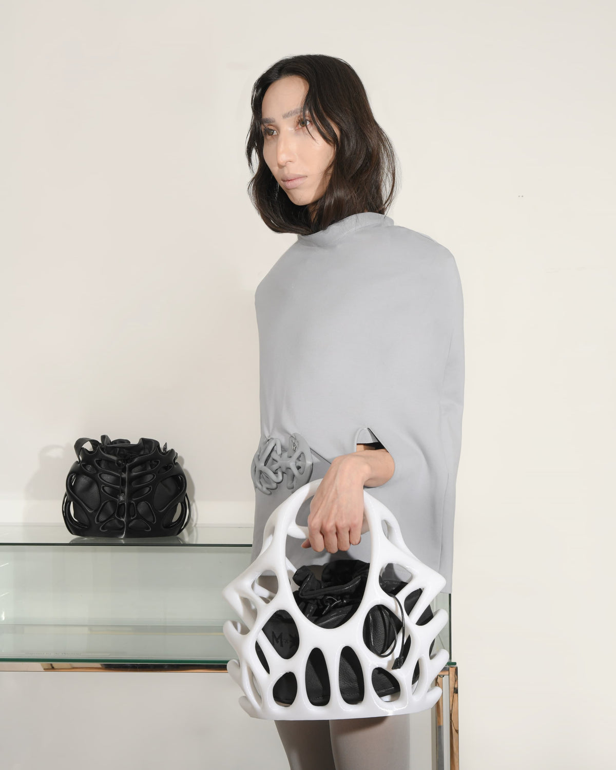 Woman holding a white handbag with black patterns, standing next to a glass table with a black bag on it.