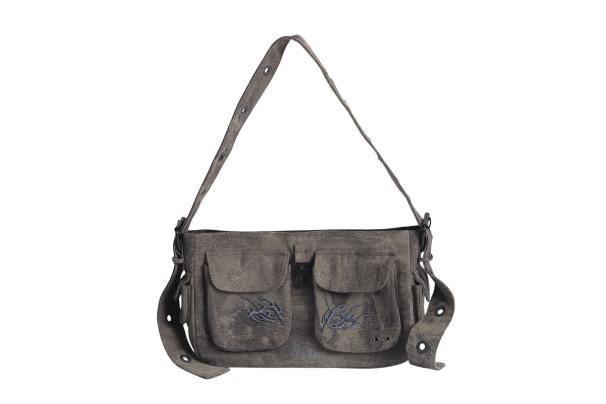 A dark gray canvas shoulder bag with a rugged, worn look, featuring two front pockets with flap closures and black embroidery designs on each pocket. The bag has an adjustable strap with metal eyelets and buckle details. A small black leather attachment hangs from one side of the bag, adding to its utilitarian aesthetic.