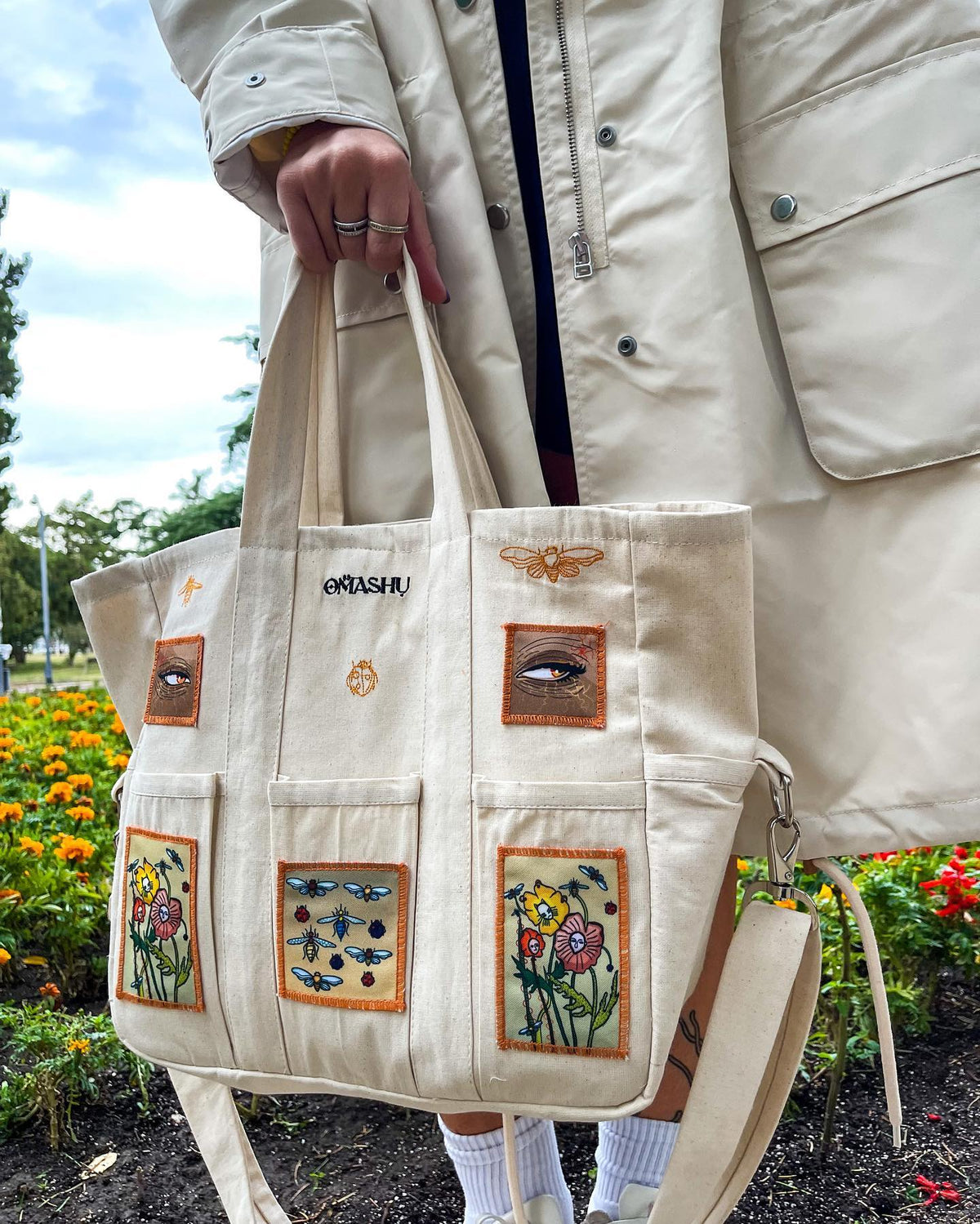 Beige tote bag with colorful patches sewn onto it. The patches depict different objects such as a bee, a flower, a butterfly, and a bird. The bag has two handles and a shoulder strap.